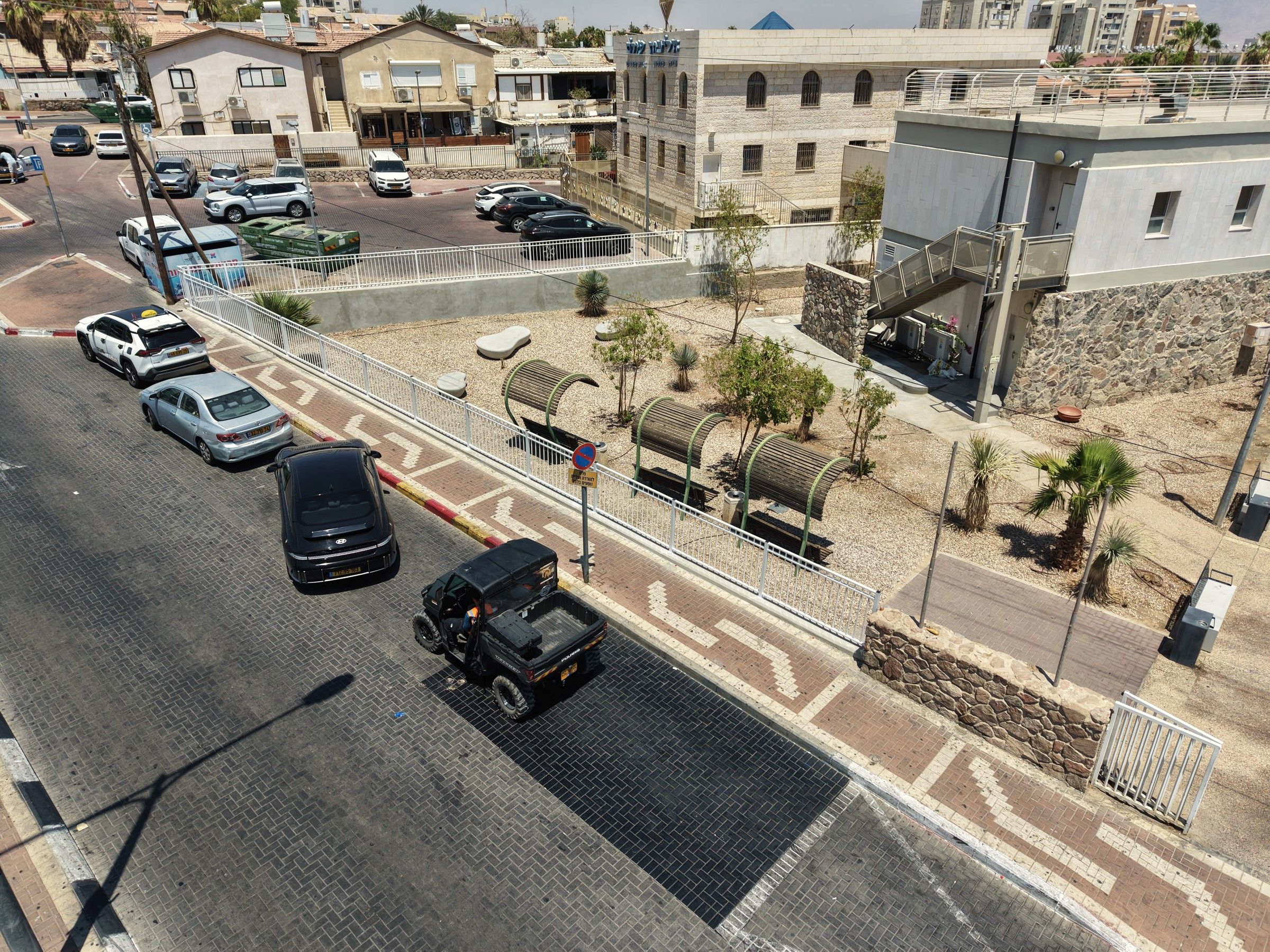 רחבה מרוצפת עם ספסלים מוצלים, צמחייה ומעקה לבן לאורך רחוב אילות
