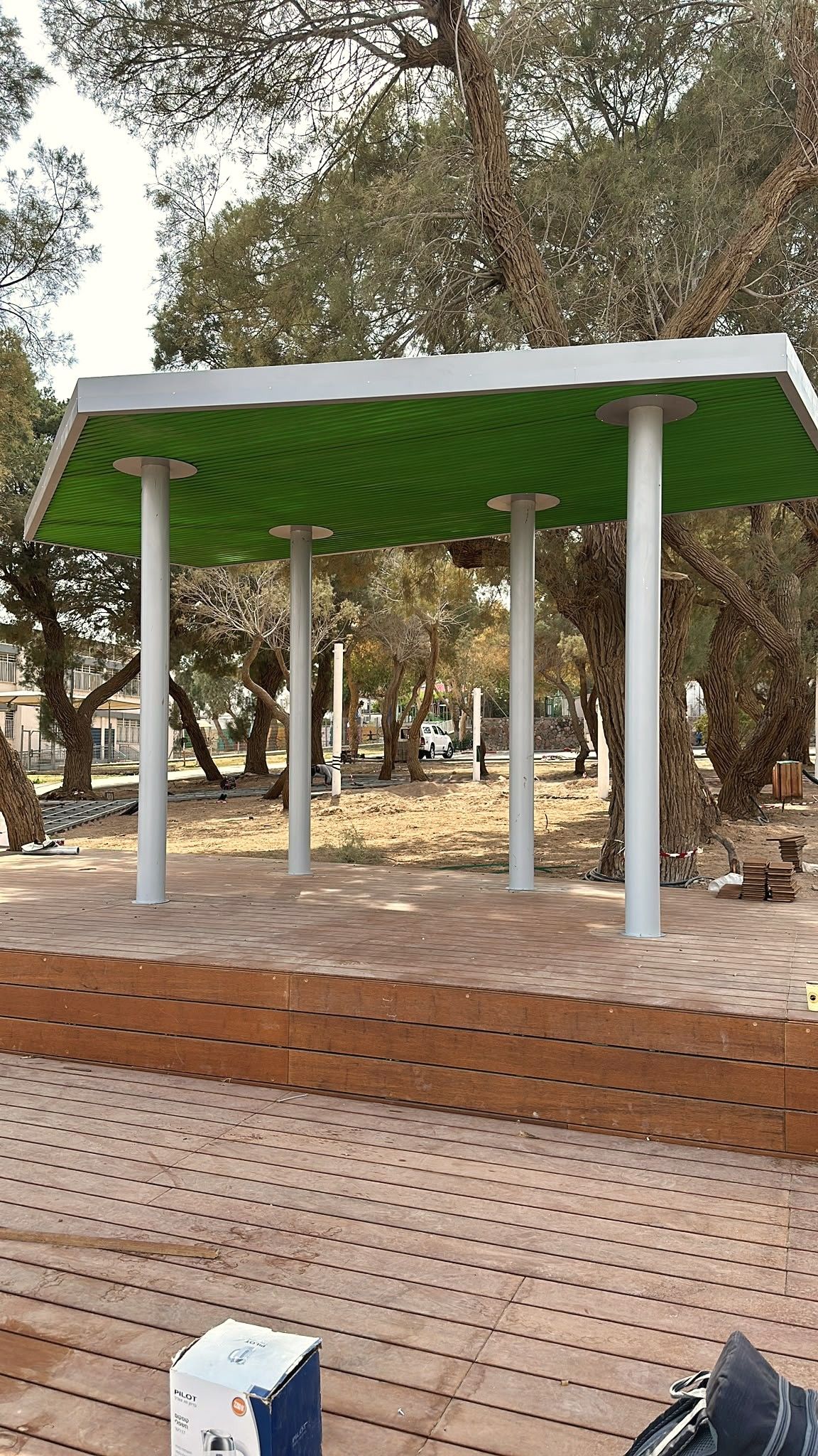 סככת הצללה עם גג ירוק על עמודי פלדה עגולים, מעל במת עץ בפארק