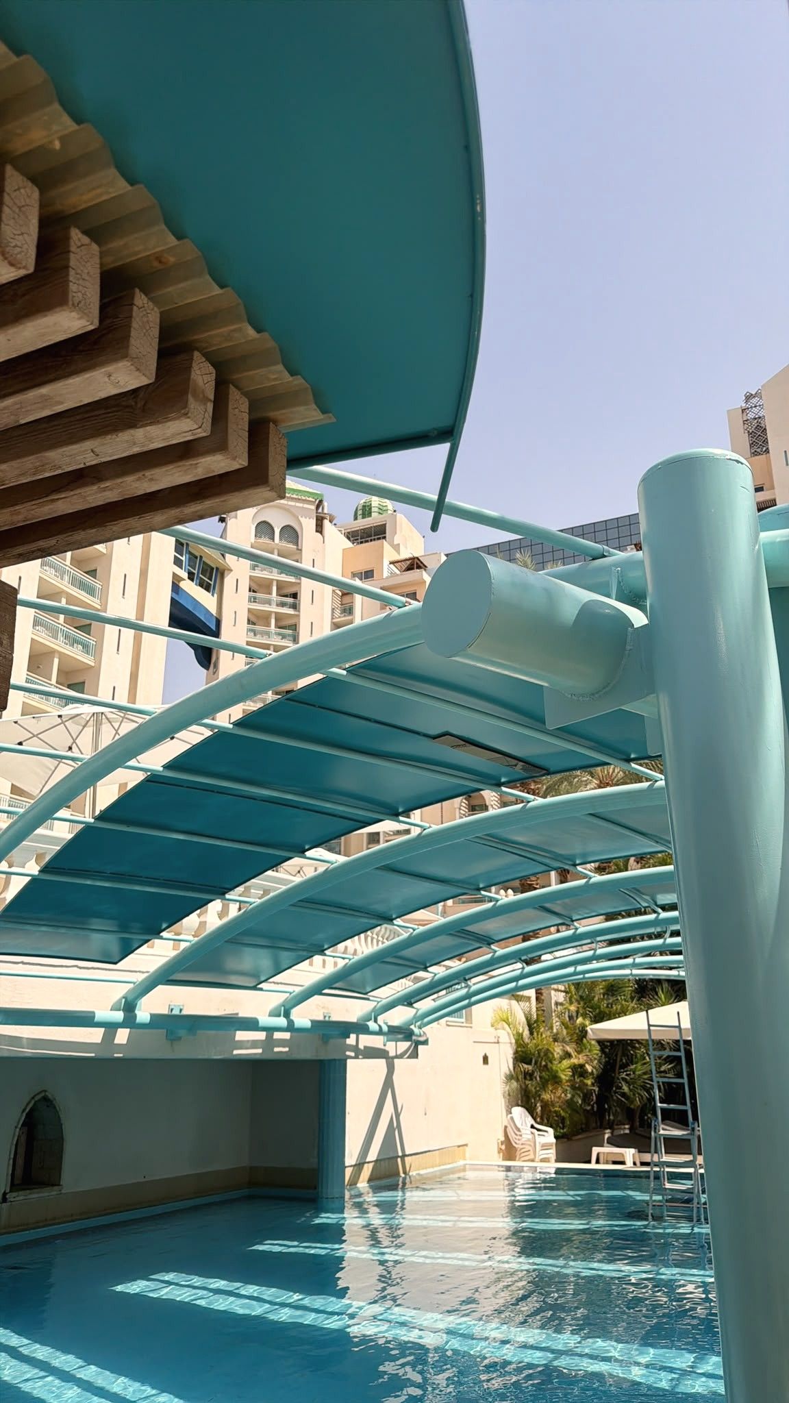 קשתות פלדה בטורקיז נושאות יריעות הצללה מעל בריכה