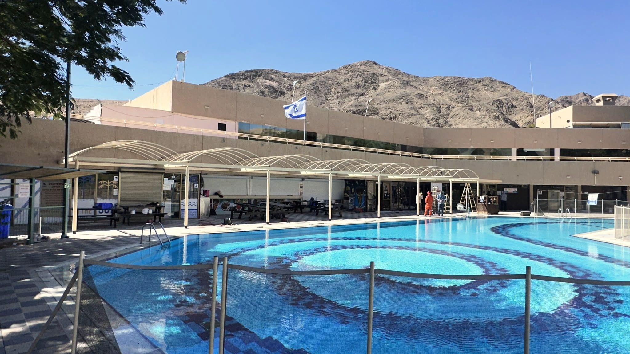 קשתות הצללה מפלדה לאורך בריכה, והרי אילת ברקע