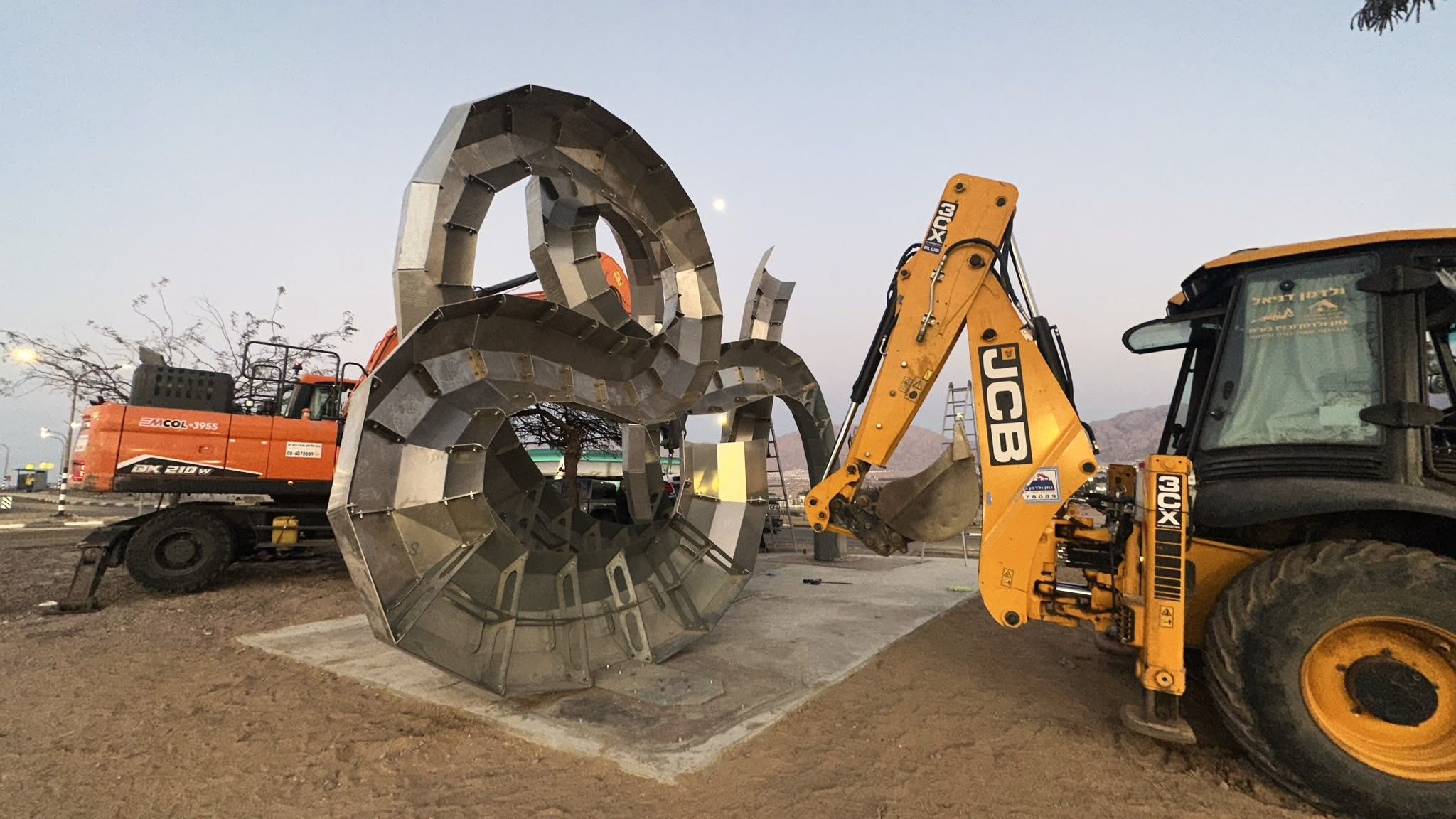 פסל נירוסטה גדול בצורת סרט מתפתל בזמן ההצבה, ומחפרון לידו