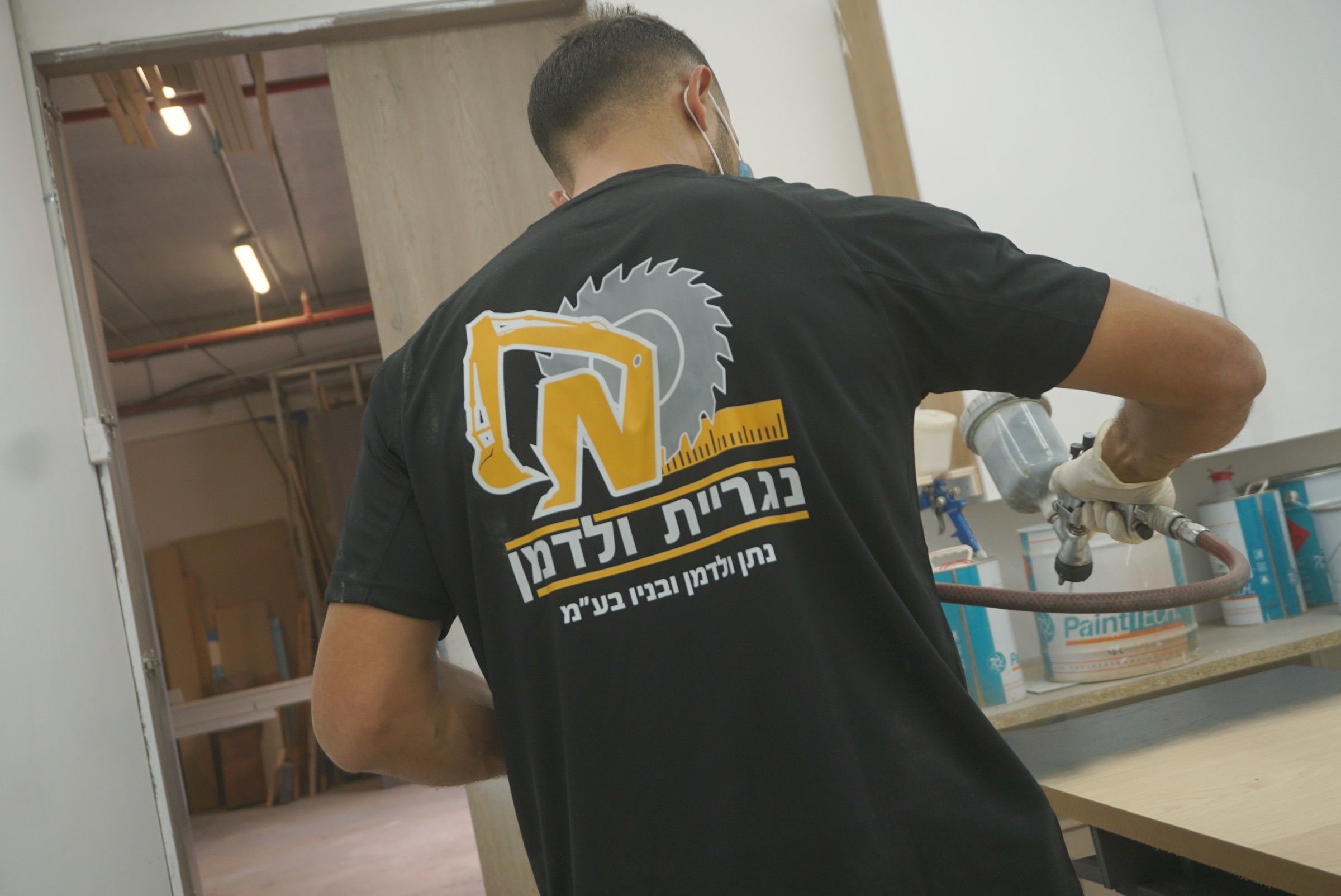 נגר בחולצת נגריית ולדמן מרסס לכה בתא הצבע של הנגרייה